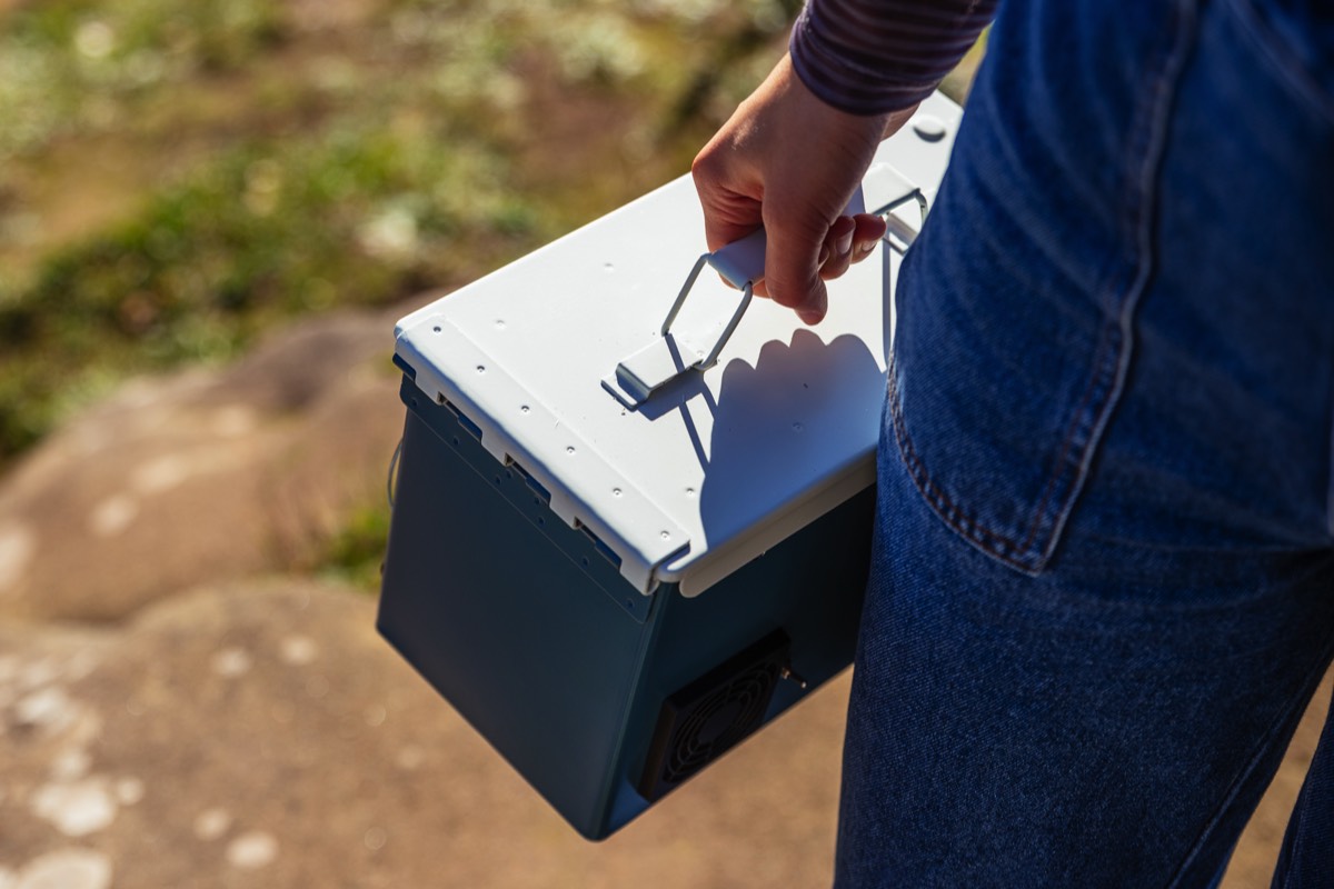 A blue and white custom pained ammo box speaker being carried in the sun